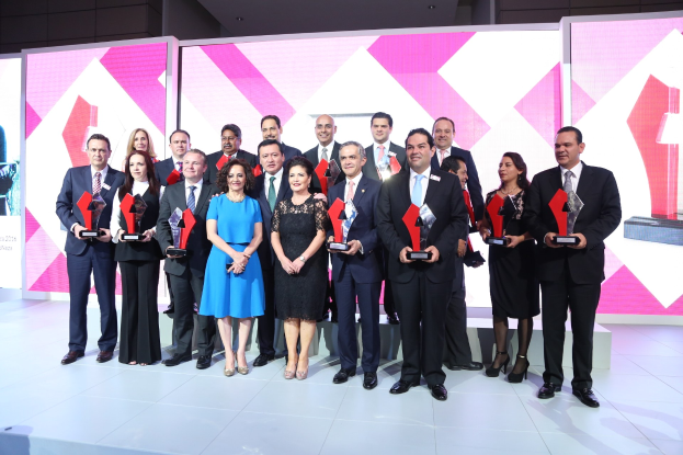 Gruppe von Menschen auf einer Bühne, die Anzüge tragen und Auszeichnungen halten, mit Bildschirmen und einer Wand im Hintergrund.