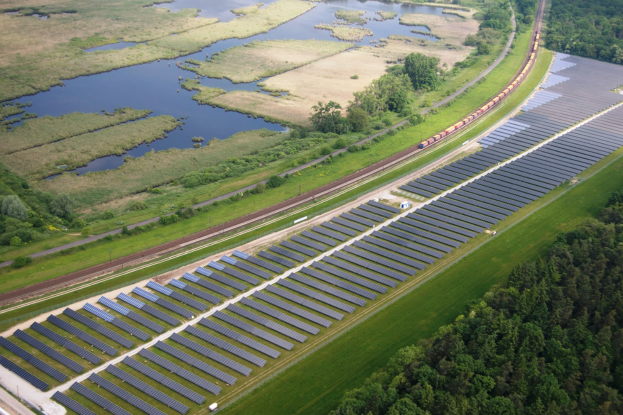 Luftaufnahme eines Solarparks mit Solarpanelen auf einem Feld, umgeben von Bäumen, Gras, Pflanzen und Wasser, mit einem Zug auf einem Bahngleis.