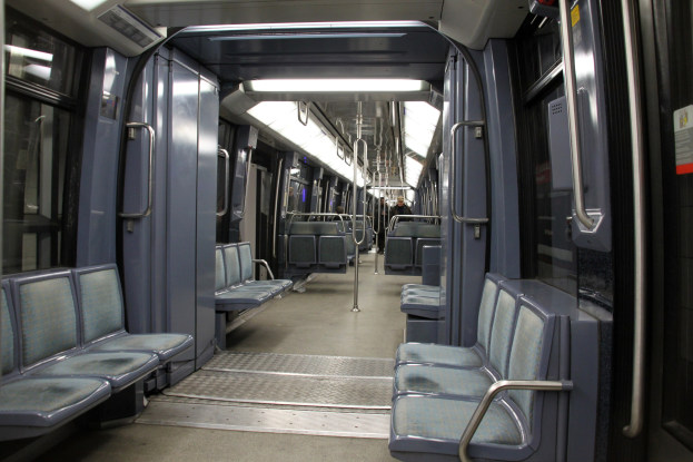 Innenseite eines U-Bahn-Wagens mit blauen Sitzen, Türen, Griffen, Stäben, Lichtern und anderen Gegenständen, mit einigen Passagieren in einer belebten Szenerie.