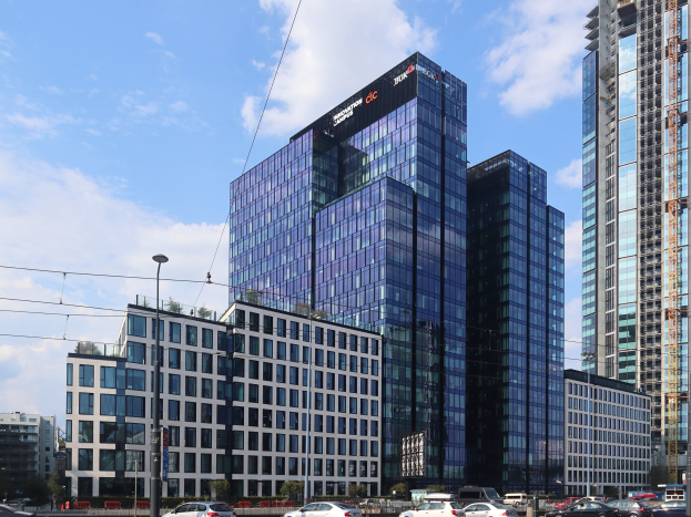 Zentrale der Deutschen Bank in Frankfurt, Deutschland, ein großes Gebäude mit vielen Fenstern, umgeben von städtischen Elementen wie Straßenlaternen, Fahrzeugen und Bäumen unter einem bewölkten Himmel.