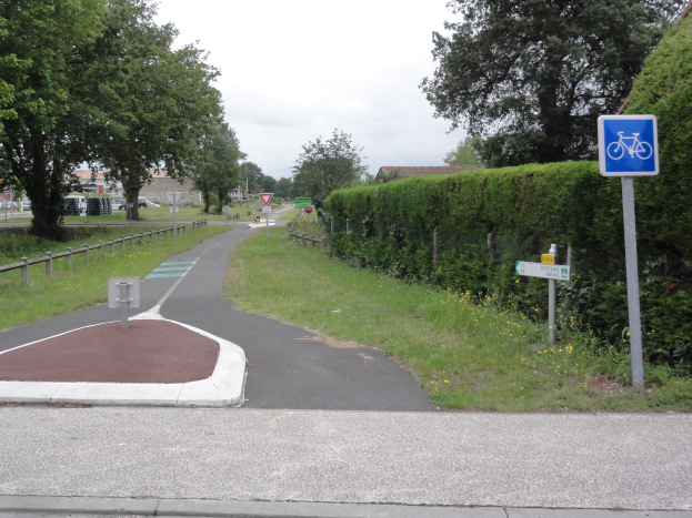 Eine Straße mit einem Fahrradweg am Rand, umgeben von Gras, Pflanzen, Bäumen und einem Zaun, mit Schildern, Fahrzeugen, Gebäuden und einem klaren blauen Himmel im Hintergrund.