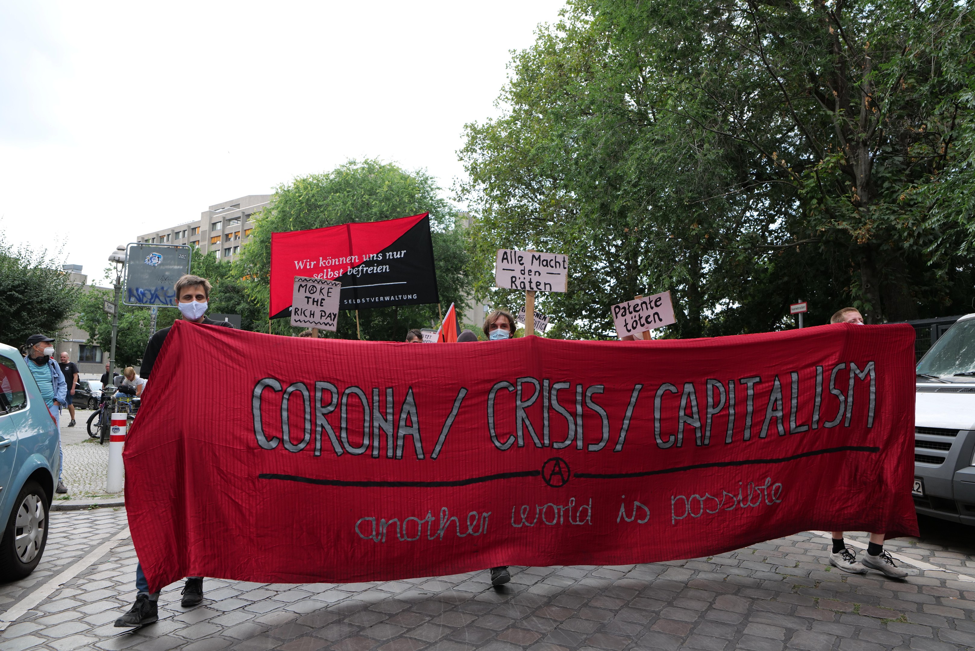 Eine Gruppe von Menschen geht die Straße entlang und trägt eine rote Fahne mit der Aufschrift "Corona-Krise/Kapitalismus", mit geparkten Fahrzeugen auf beiden Seiten und Gebäuden, Bäumen und einem klaren Himmel im Hintergrund.